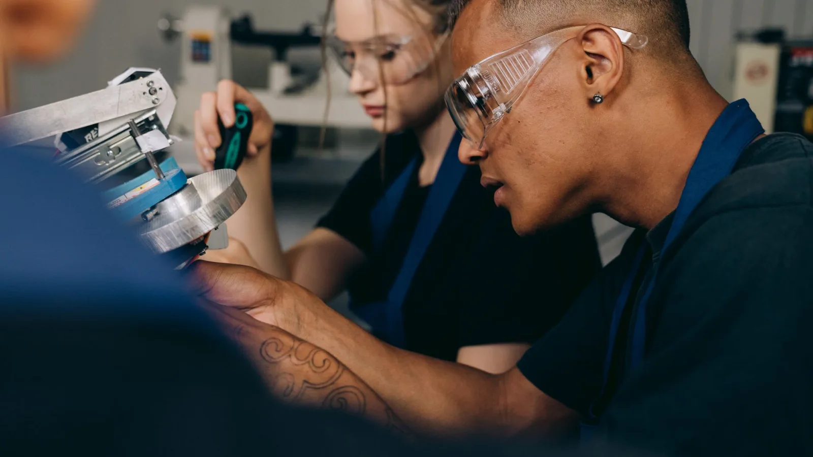Two young adults working attentively in a workshop, wearing protective goggles.