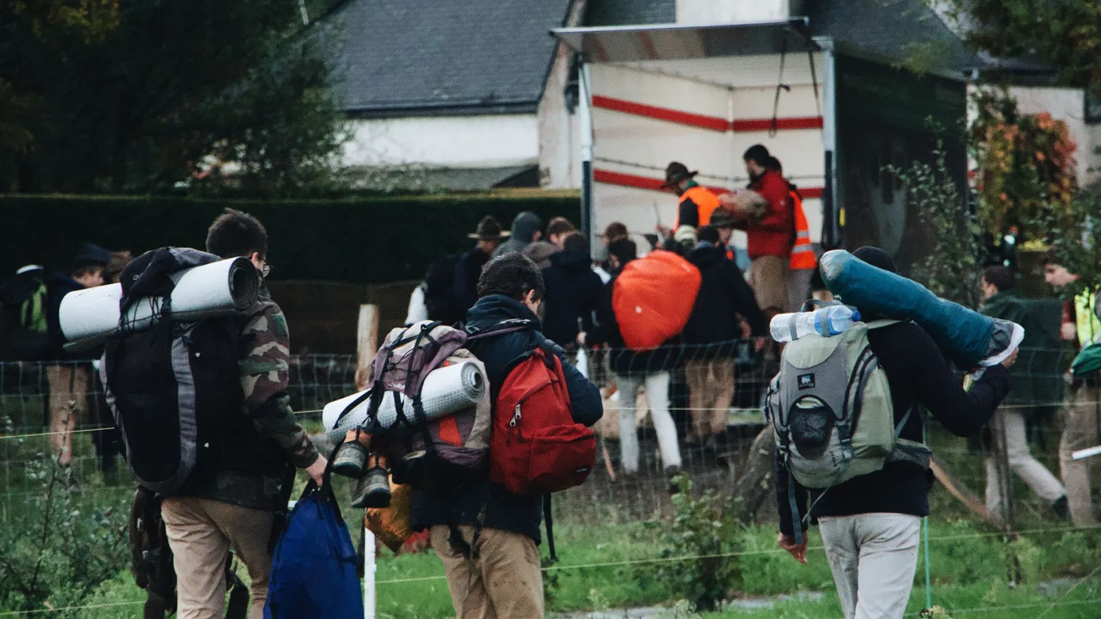 A group of people carrying backpacks and camping gear heading to a camp truck in a green outdoor setting.