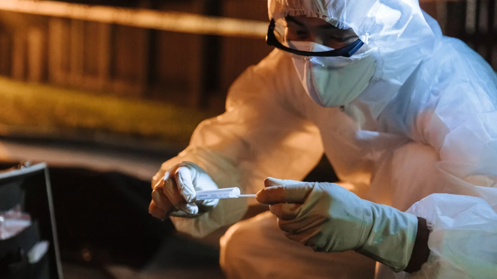 A forensic investigator in protective gear collects evidence outdoors at night.