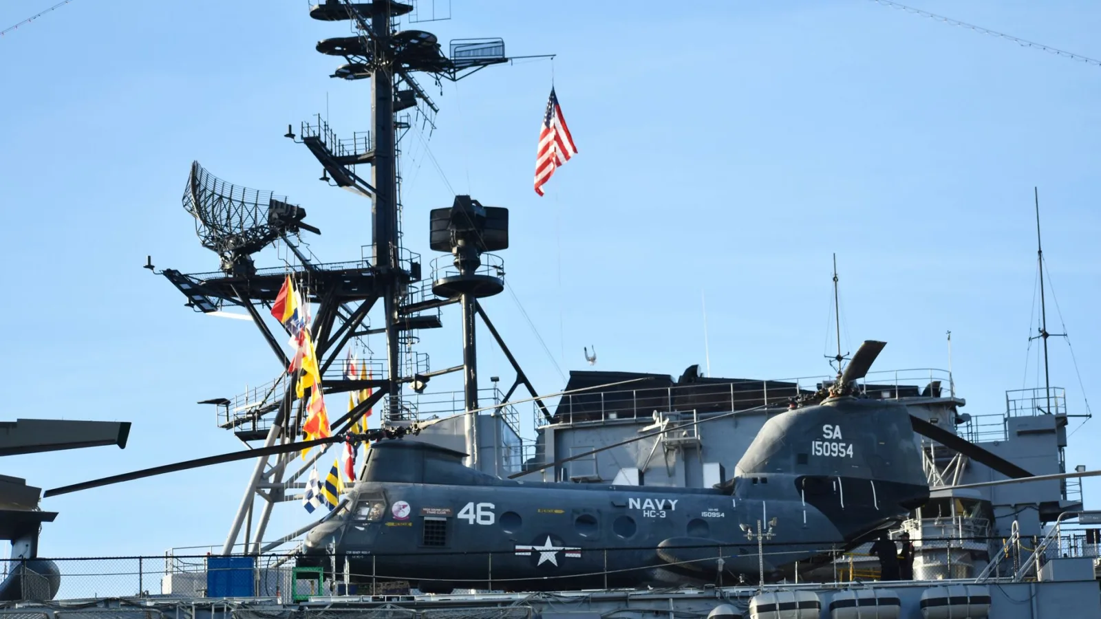 A US Navy helicopter is stationed on the deck of an aircraft carrier with radar equipment in the background.