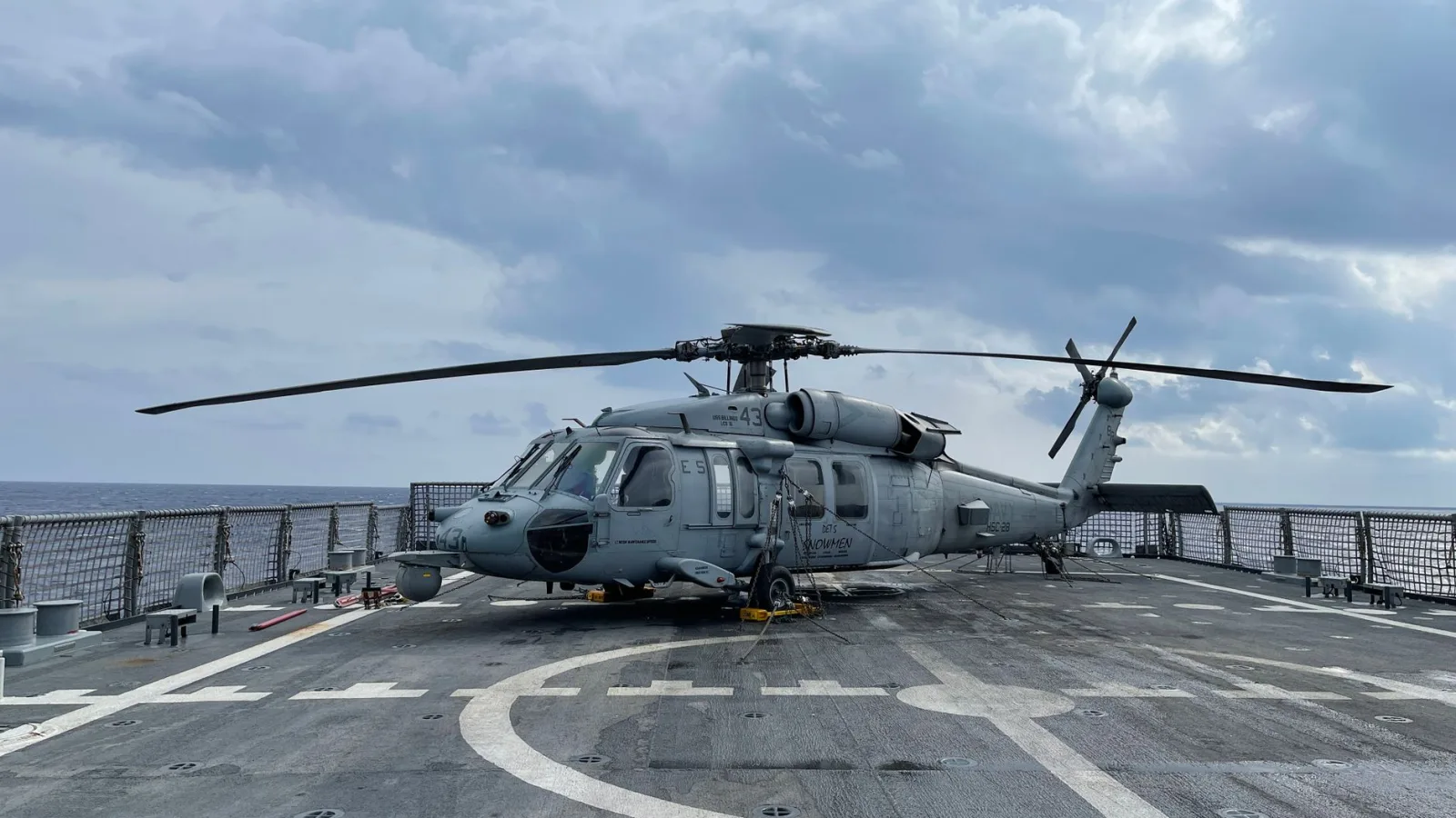 A military helicopter on a naval flight deck under a cloudy sky at sea.